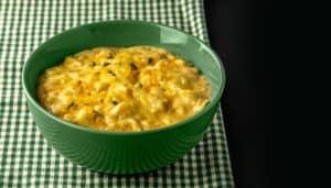 Green bowl filled with creamy macaroni and cheese garnished with herbs, placed on a green and white checkered cloth.