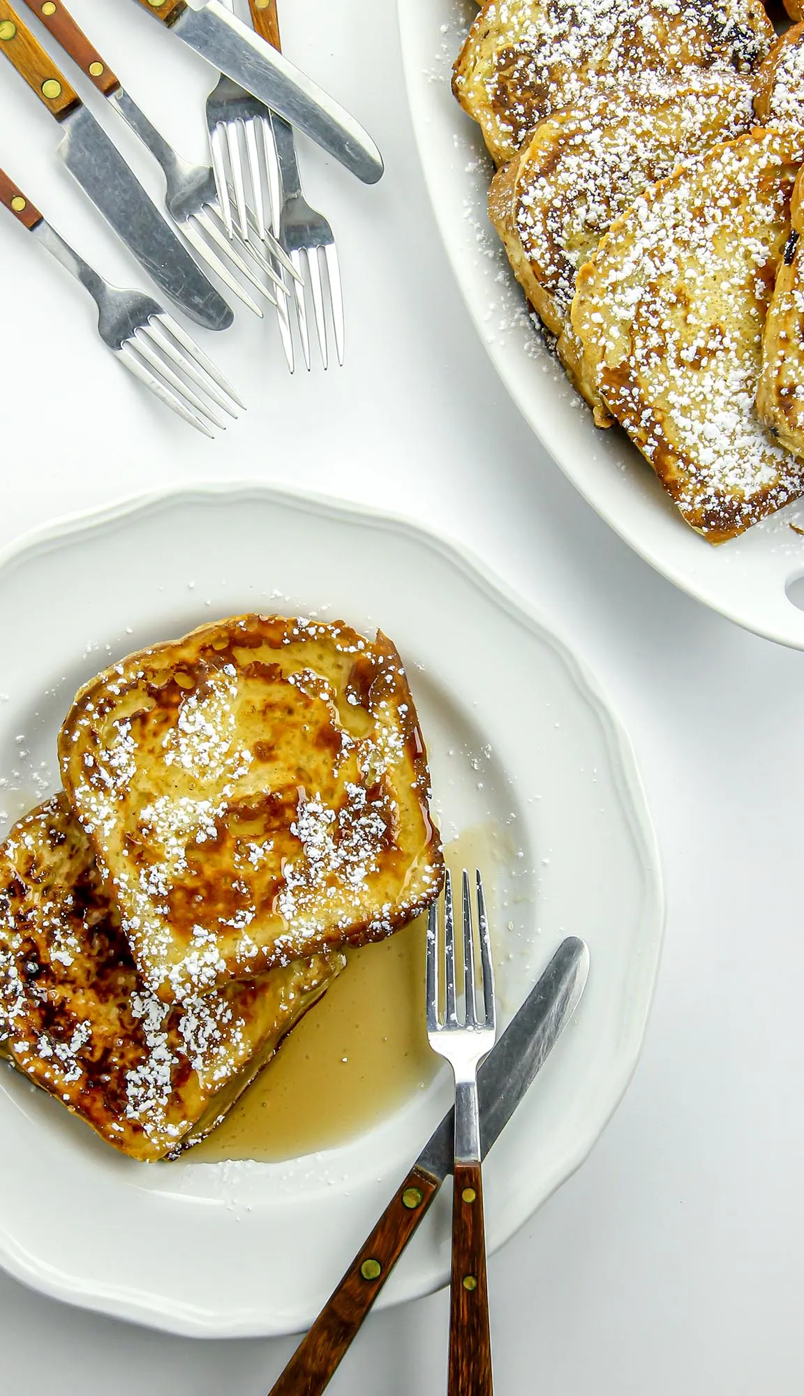 Two slices of French toast with powdered sugar and syrup on a white plate, with forks and knives nearby.
