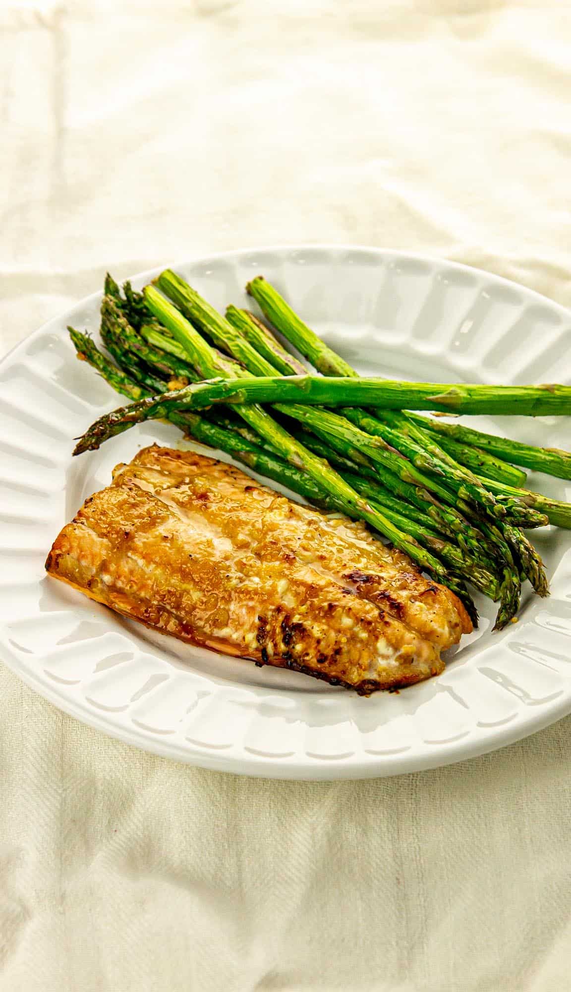 Grilled salmon fillet and roasted asparagus spears served on a white plate with a light cloth background.