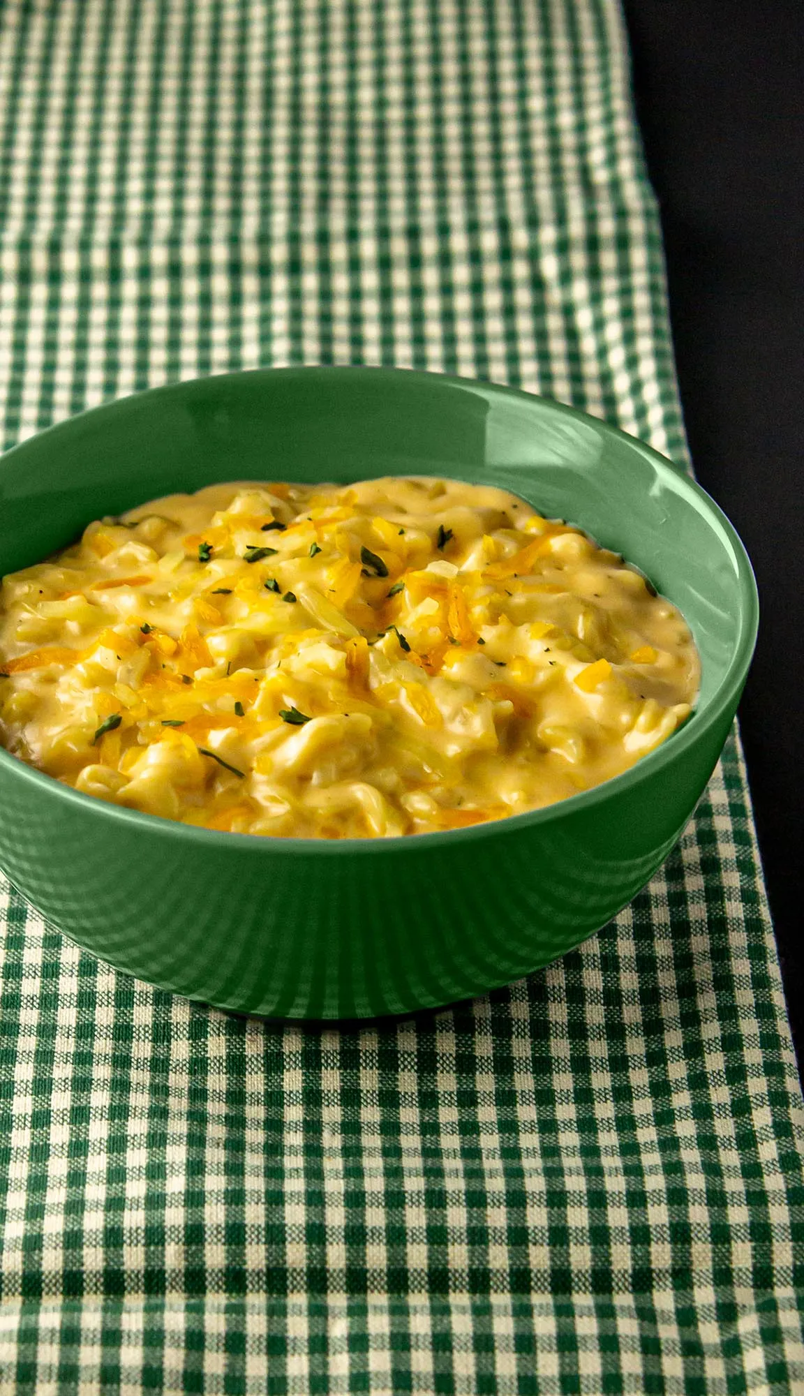 Green bowl filled with creamy cheddar macaroni garnished with herbs on a green and white checkered cloth.
