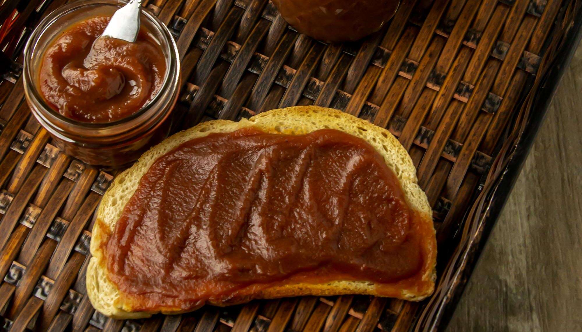 Slice of bread spread with a thick layer of brown fruit butter, next to a jar filled with the same fruit butter and a white spreading knife.