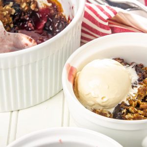 Berry crisp topped with a scoop of vanilla ice cream in a white bowl, next to a larger white dish of berry crisp with a spoon.