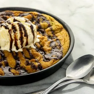 Large chocolate chip cookie topped with vanilla ice cream and chocolate syrup, served in a pan with two spoons on a marble surface.