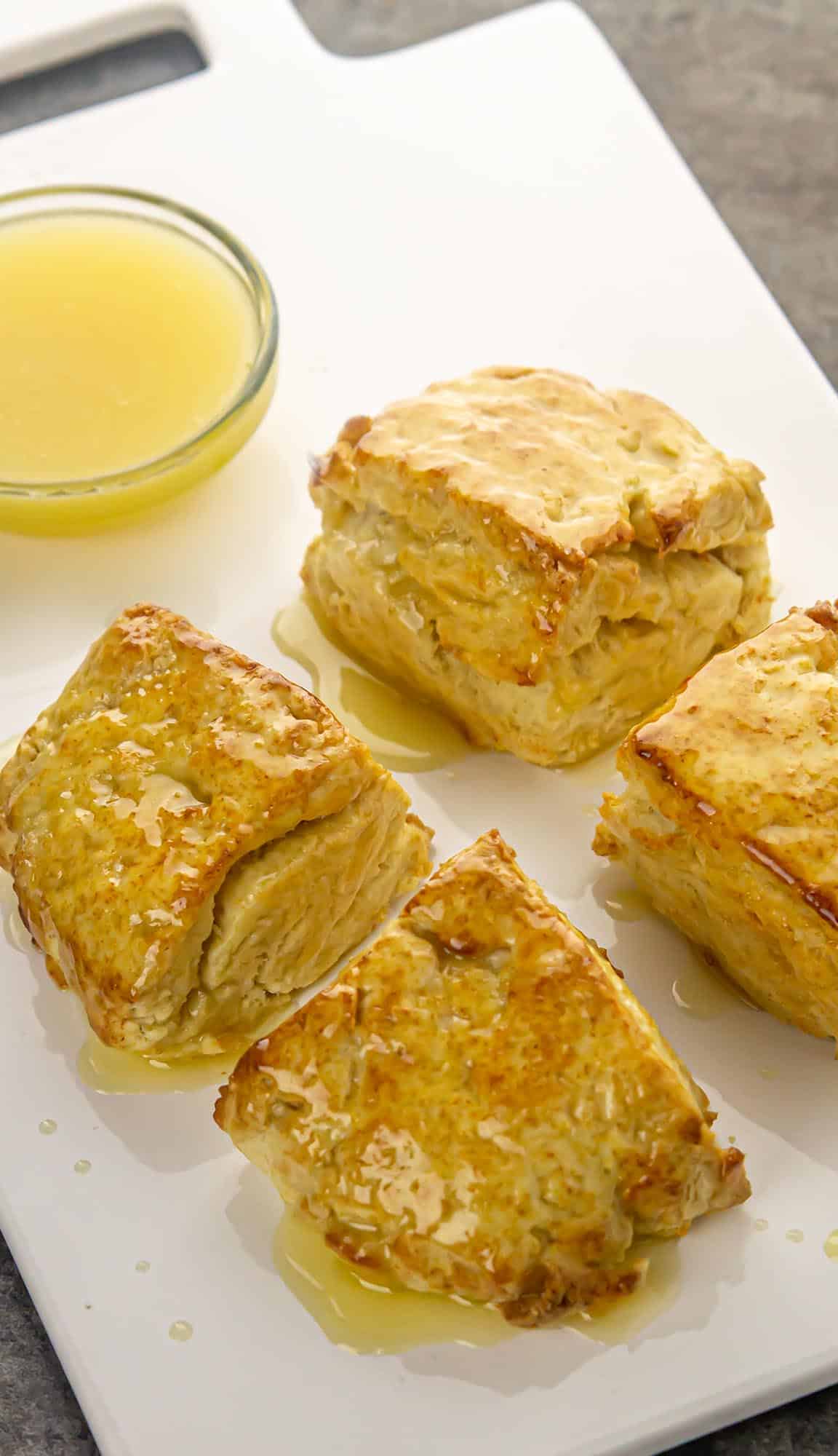 Four golden scones drizzled with syrup on a white tray with a small bowl of extra syrup.