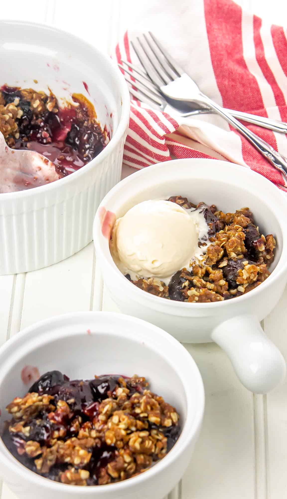 Three white ramekins with dark berry crisp, one topped with a scoop of vanilla ice cream, on a white table with a red-striped cloth and two forks.