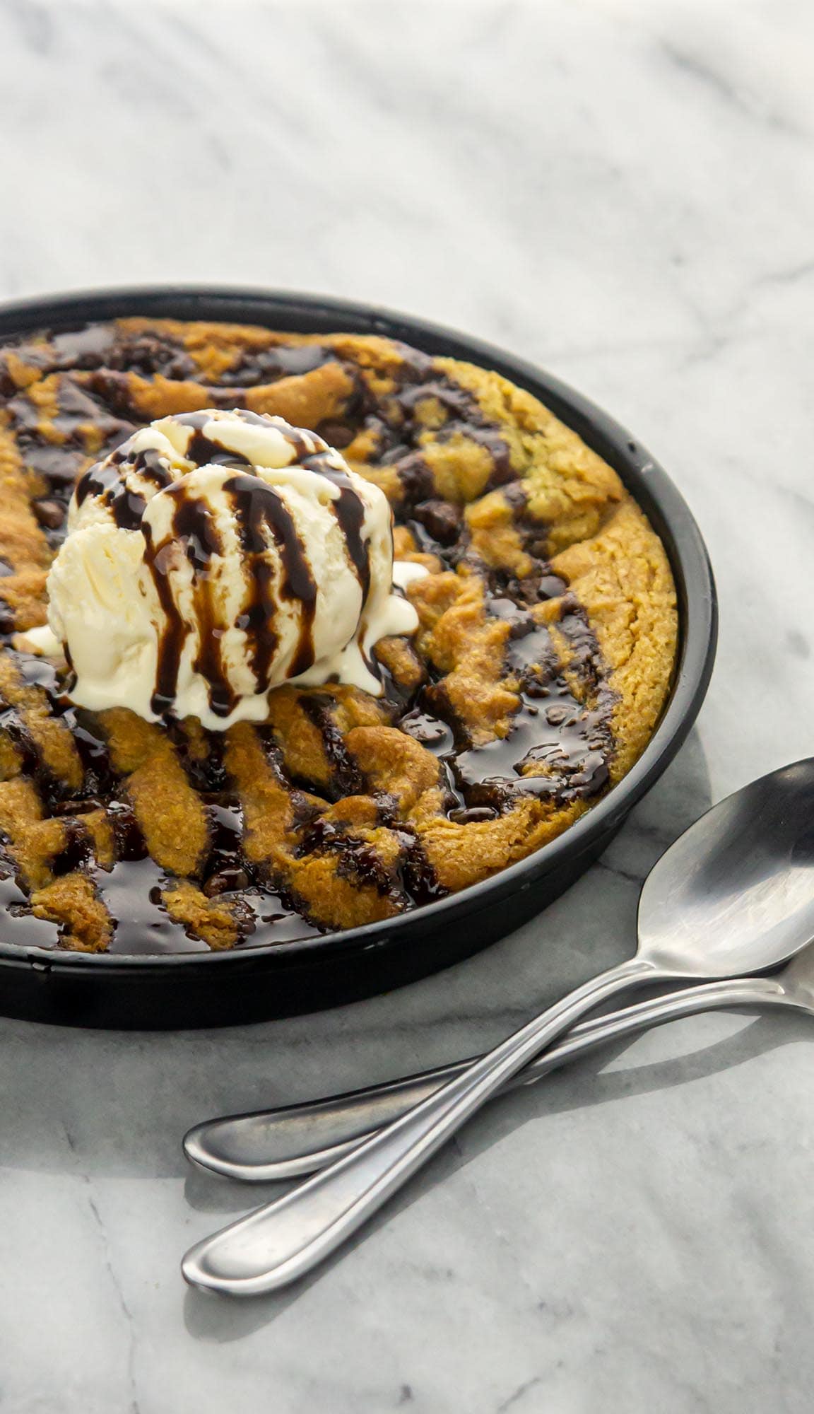 Chocolate chip skillet cookie topped with vanilla ice cream and chocolate syrup, served with two spoons on a marble surface.