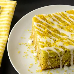 Square piece of yellow cake with white icing drizzle and yellow sugar sprinkles on white plate, next to two forks on a yellow striped napkin.