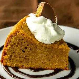 Slice of pumpkin cake with whipped cream and a peanut butter cup on top, served on a plate with chocolate syrup swirl.