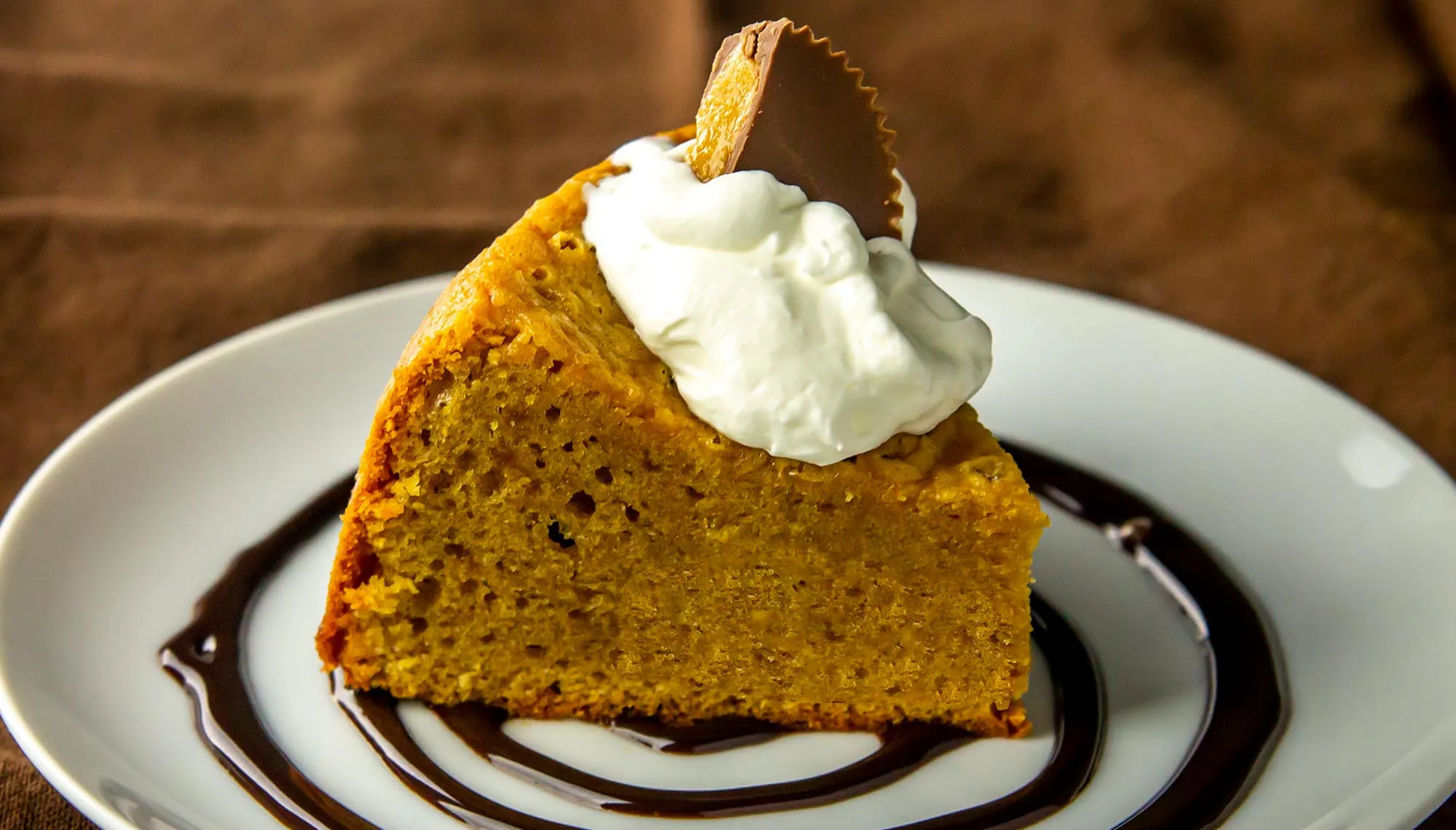 Slice of pumpkin cake with whipped cream and a peanut butter cup on top, served on a plate with chocolate syrup swirl.