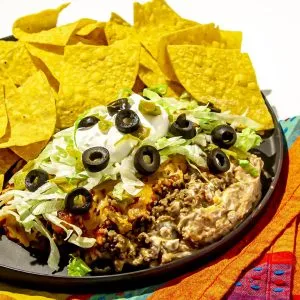 Ground beef and bean dip topped with shredded lettuce, black olives, sour cream, and jalapeños served with tortilla chips on a black plate.