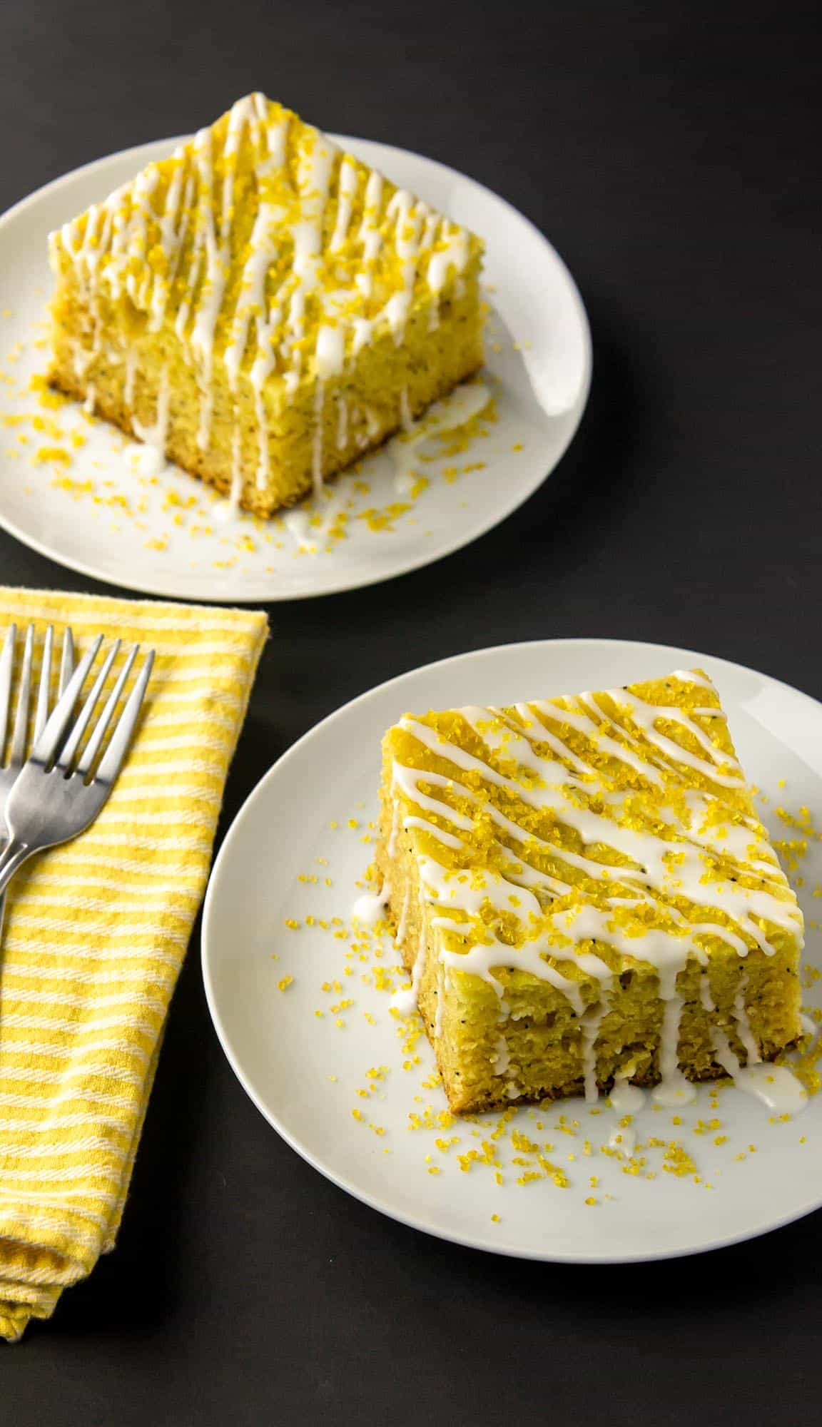 Two square pieces of lemon cake with white icing drizzle and yellow sprinkles on white plates next to yellow striped napkin and forks.