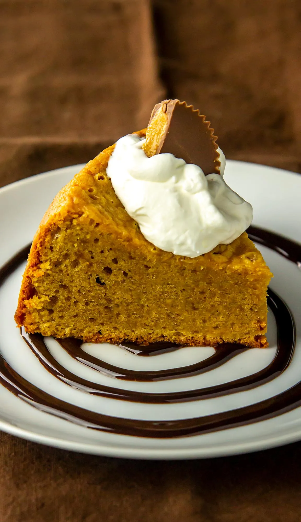 Slice of pumpkin cake topped with whipped cream and half a peanut butter cup, served on a plate with chocolate sauce spiral.