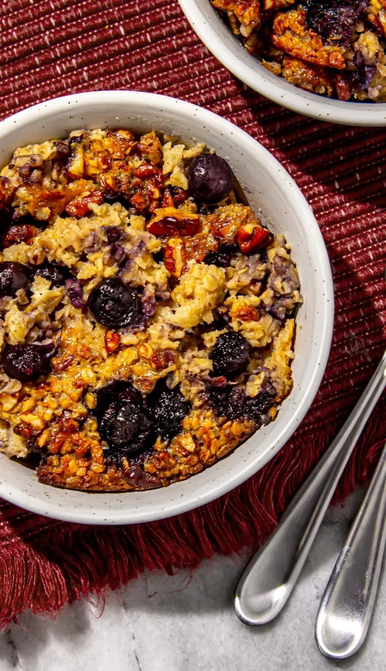 Baked oatmeal with blueberries and pecans in a white bowl on a red textured cloth with silver spoons nearby.