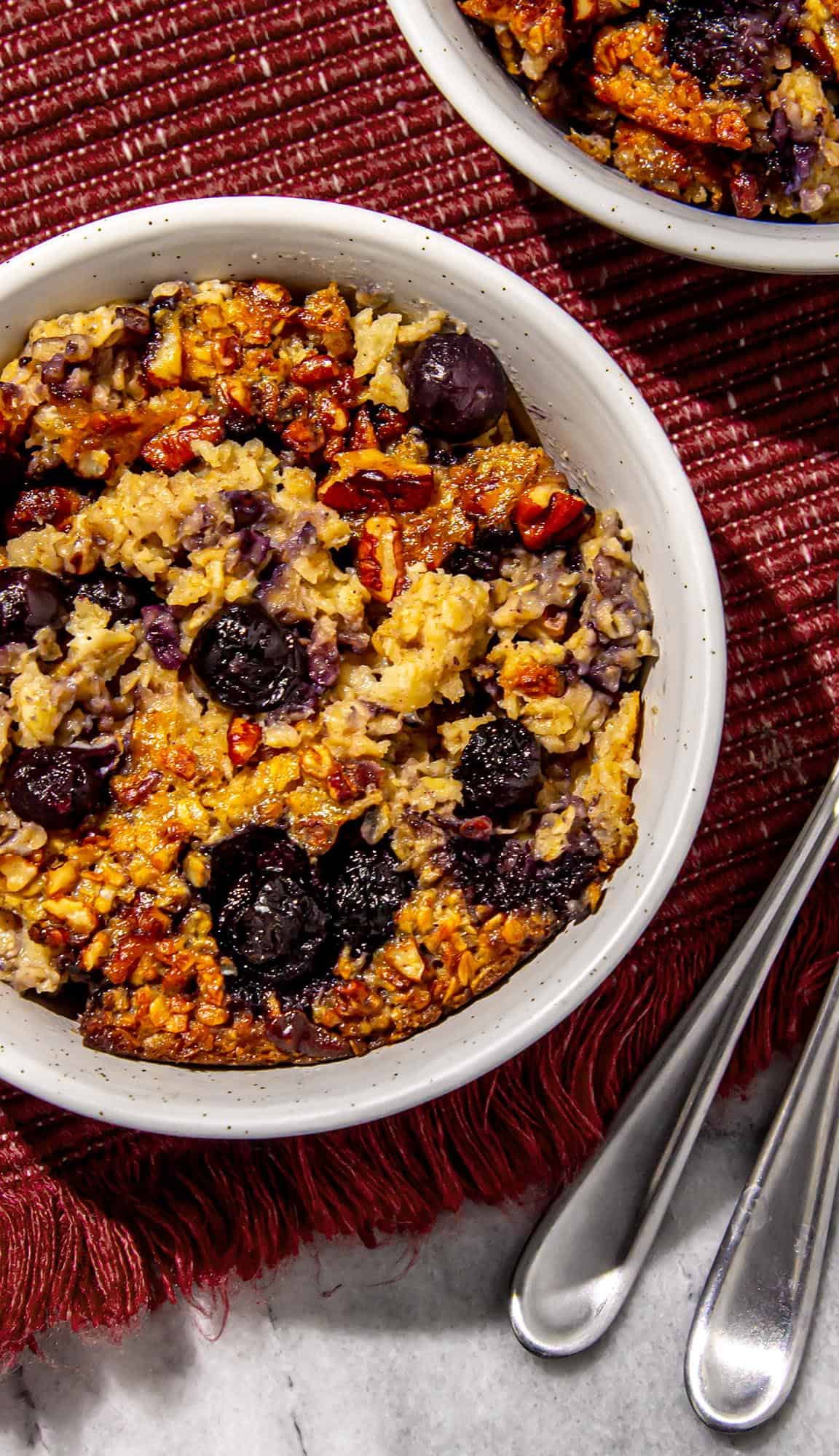 Baked oatmeal with blueberries and pecans in a white bowl on a red textured cloth with silver spoons nearby.