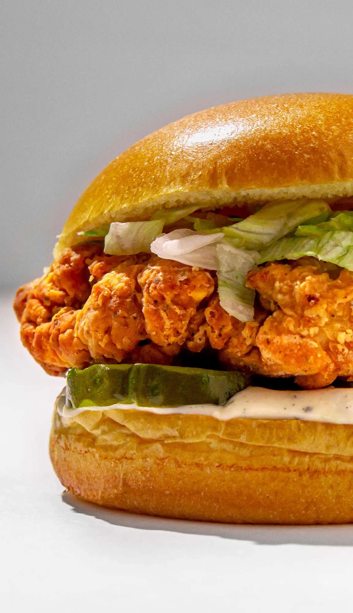 Crispy fried chicken sandwich with lettuce, pickles, and mayonnaise on a toasted bun against a plain background.