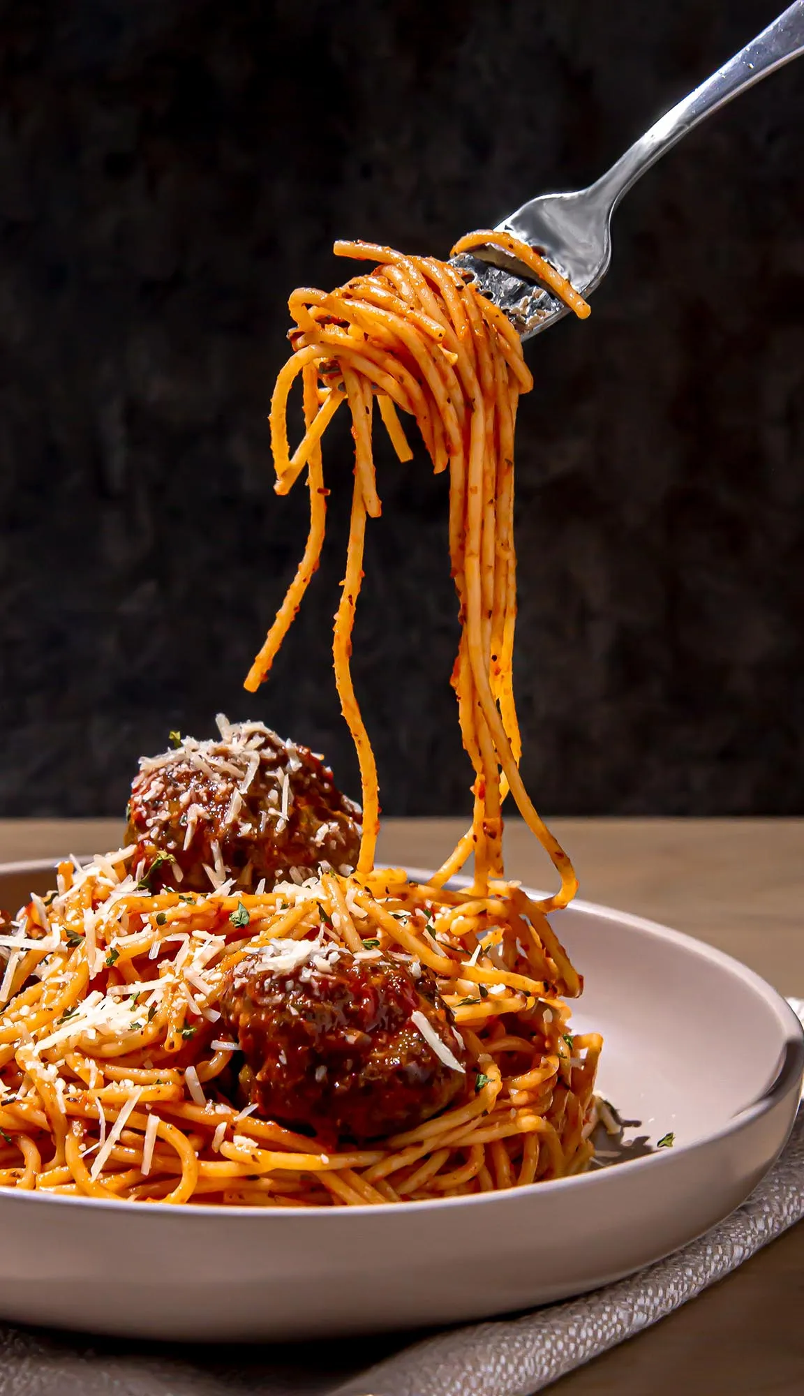 Spaghetti twirled on a fork above a plate of spaghetti and meatballs topped with grated cheese and herbs.