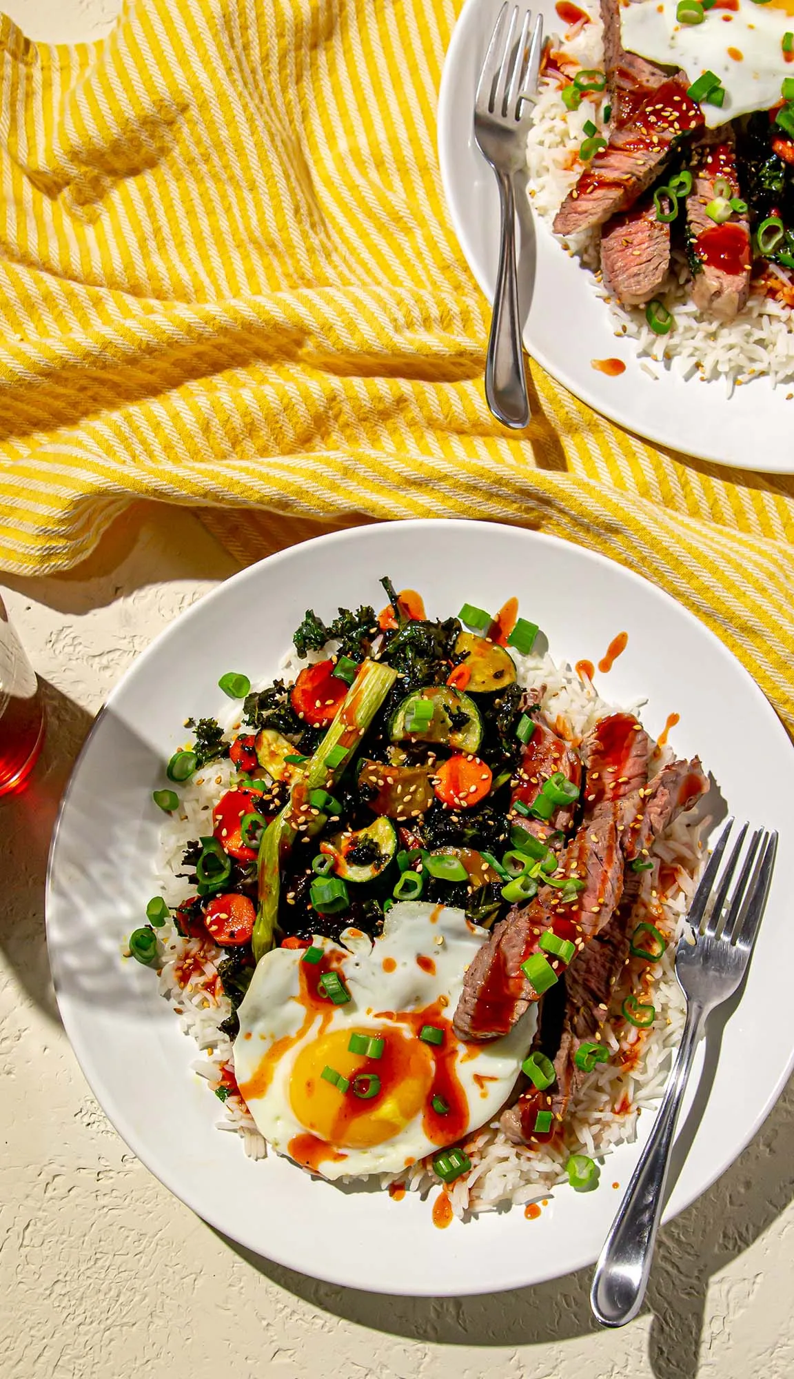 Rice bowls with fried egg, sliced steak, sautéed vegetables, green onions, sesame seeds, and hot sauce on white plates with forks on a yellow-striped cloth.