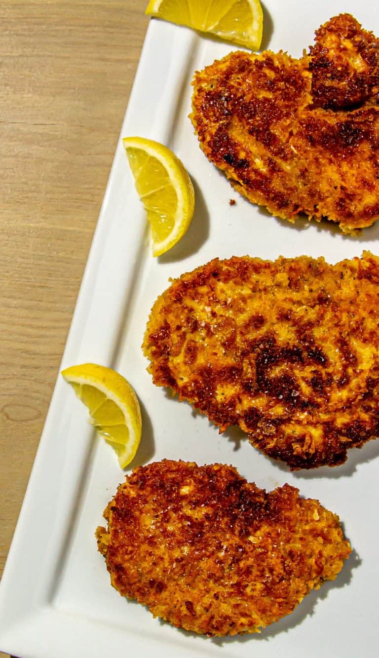 Three crispy breaded chicken cutlets on a white platter with lemon wedges on a wooden table.