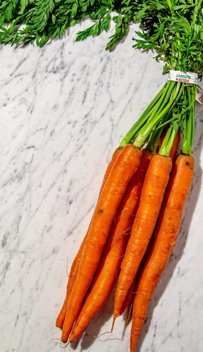 Bunch of six fresh orange carrots with green tops on a white marble surface.