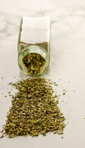 Glass spice jar tipped over with dried oregano spilled on a light surface.