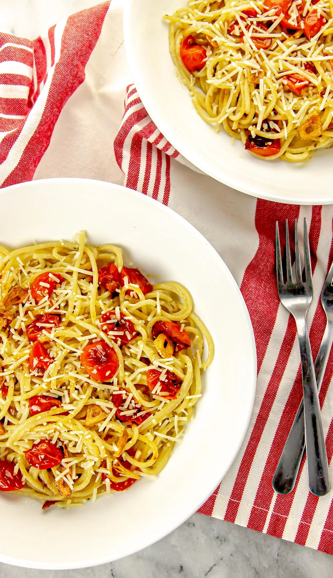 Two white bowls with spaghetti, cherry tomatoes, garlic slices, and grated cheese on white and red striped cloth with two forks.
