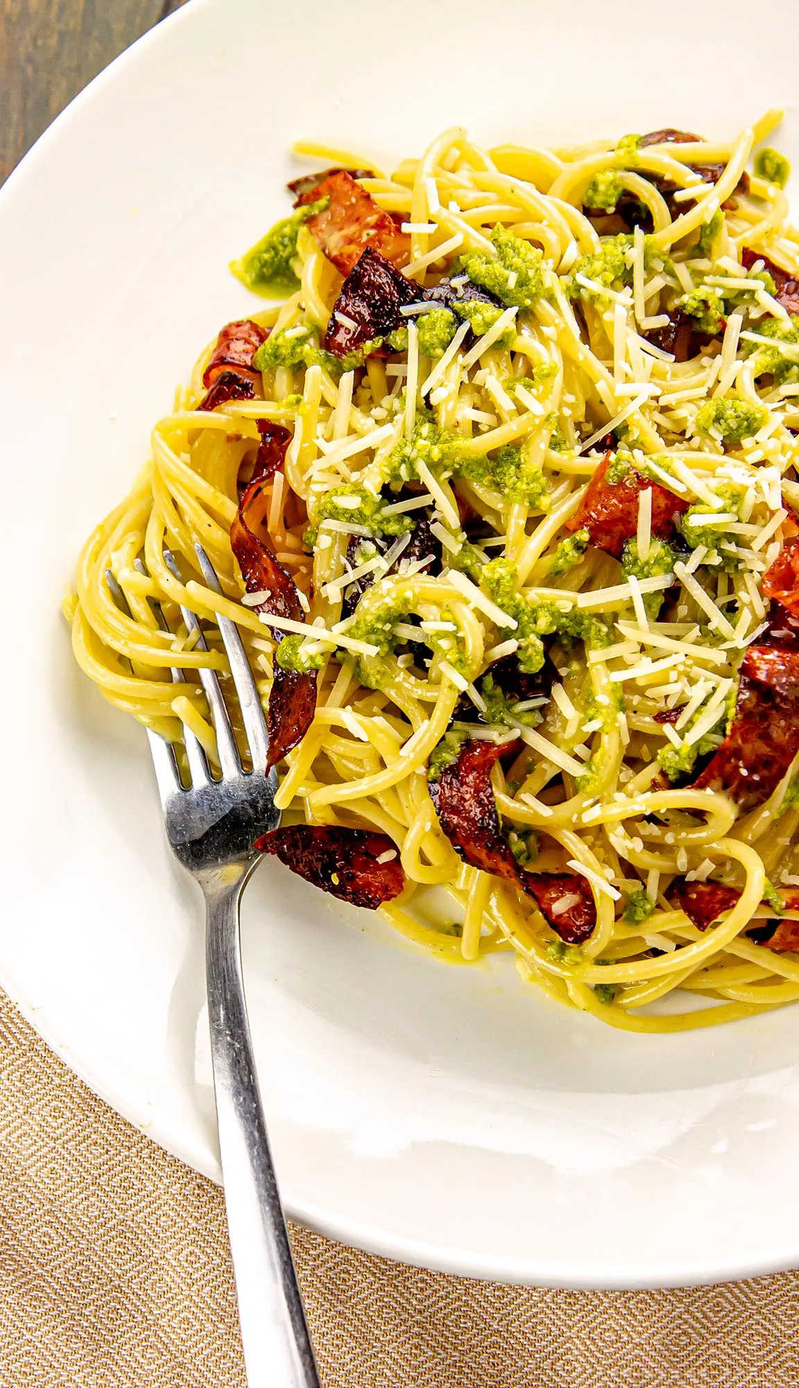 Spaghetti with green pesto sauce, grated cheese, and crispy bacon pieces on a white plate with a fork.