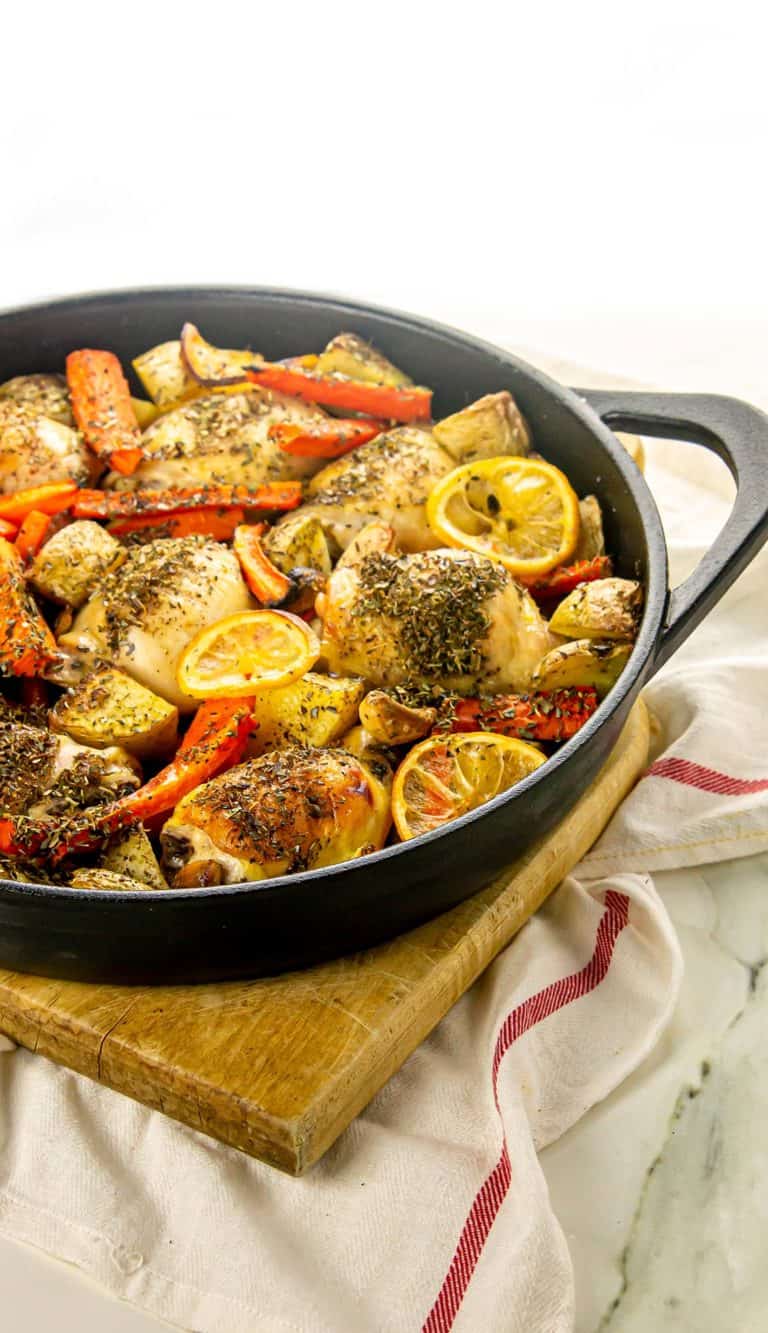 Seasoned roasted chicken thighs with lemon slices, carrots, potatoes, and mushrooms in a black skillet on a wooden board with a white towel.