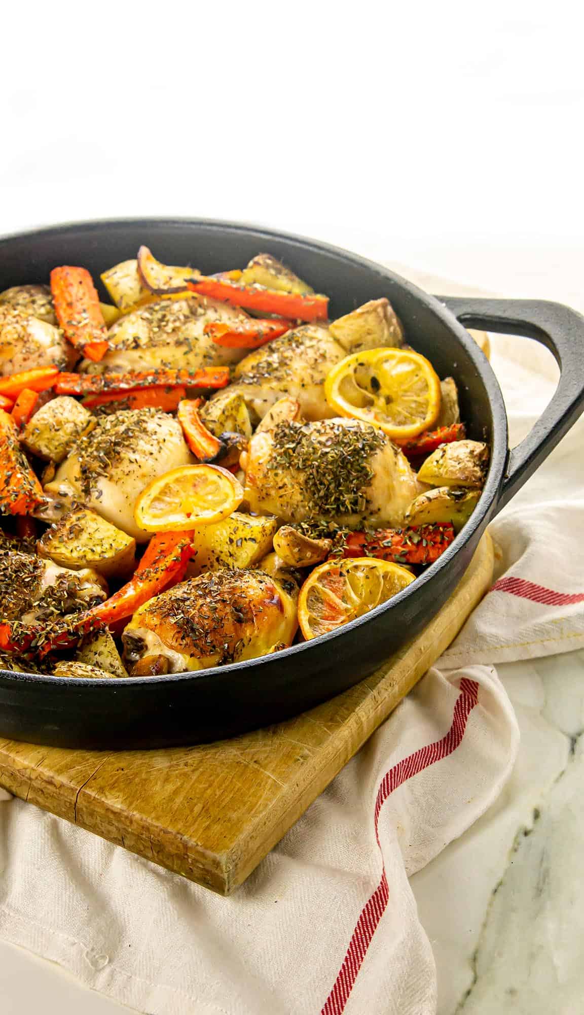 Seasoned roasted chicken thighs with lemon slices, carrots, potatoes, and mushrooms in a black skillet on a wooden board with a white towel.