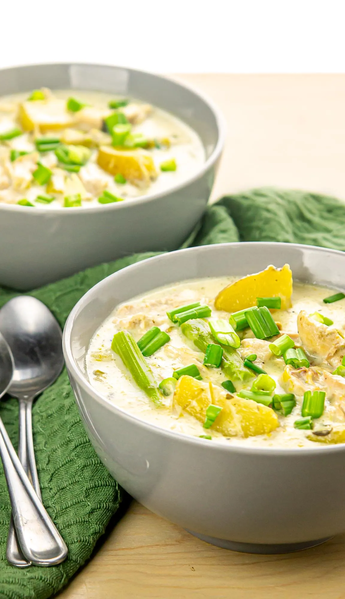 Creamy soup with asparagus, yellow squash, chicken, and chopped green onions in white bowls on a green cloth with two spoons nearby.
