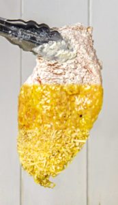 Chicken breast dipped in flour, then beaten egg, and coated with grated cheese held by tongs.