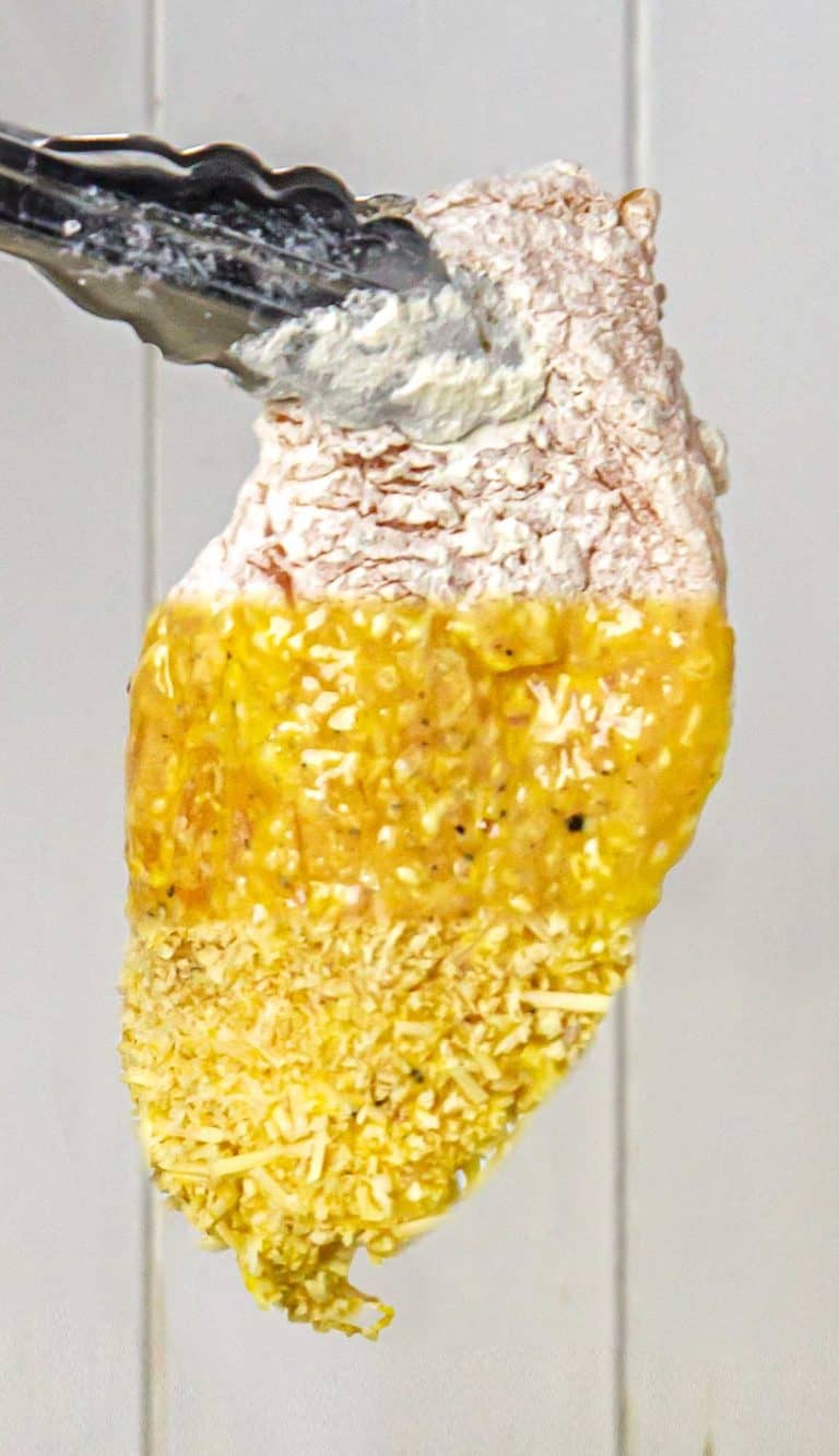 Chicken breast dipped in flour, then beaten egg, and coated with grated cheese held by tongs.