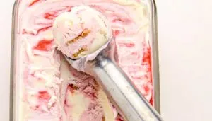 Scoop of strawberry swirl ice cream with vanilla and nut pieces in a metal scooper inside a glass container.