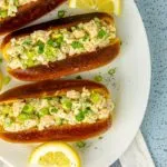 Three shrimp salad sandwiches on toasted buns garnished with chopped green onions and lemon wedges on a white plate.