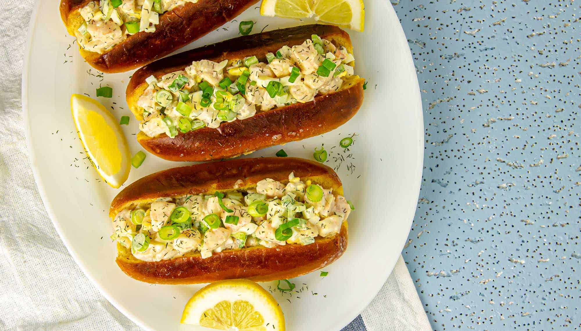 Three shrimp salad sandwiches on toasted buns garnished with chopped green onions and lemon wedges on a white plate.