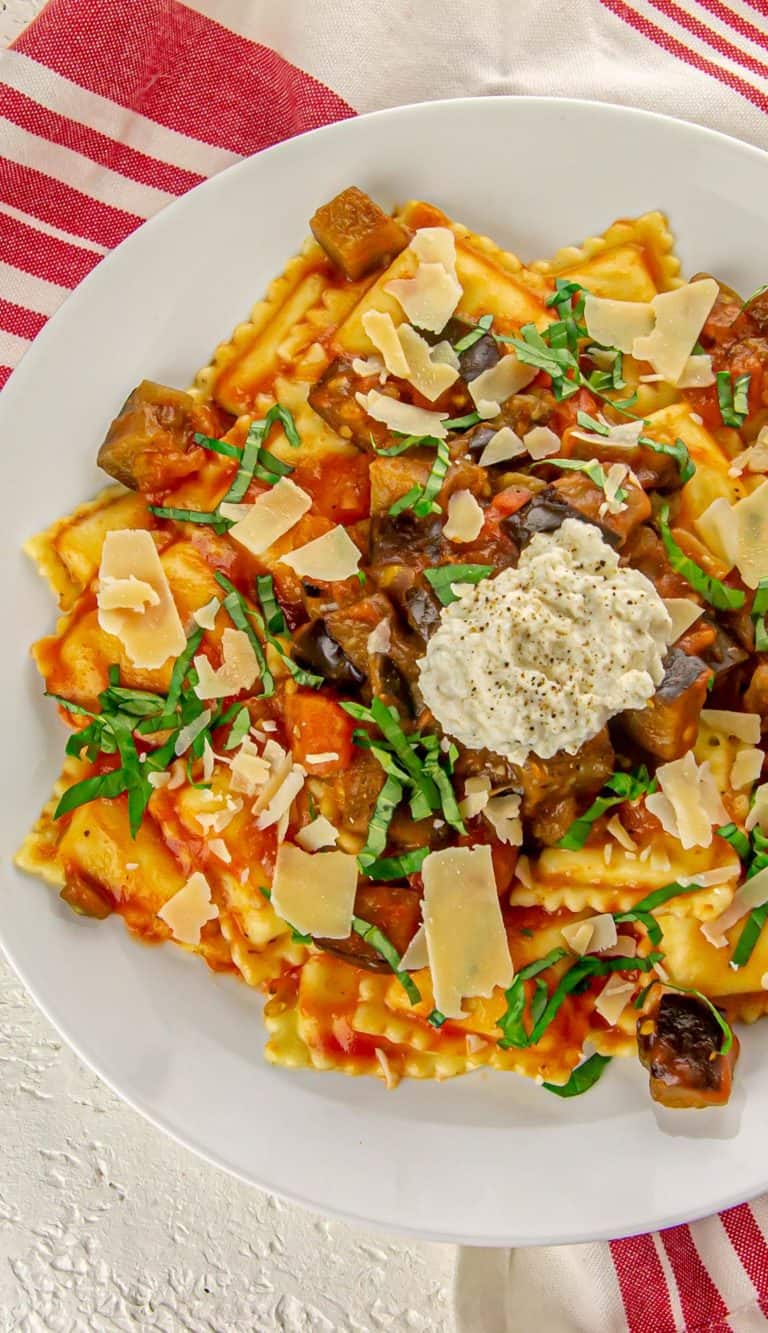 Ravioli with eggplant, tomato sauce, shaved Parmesan, chopped basil, and a dollop of ricotta sprinkled with pepper on a white plate.