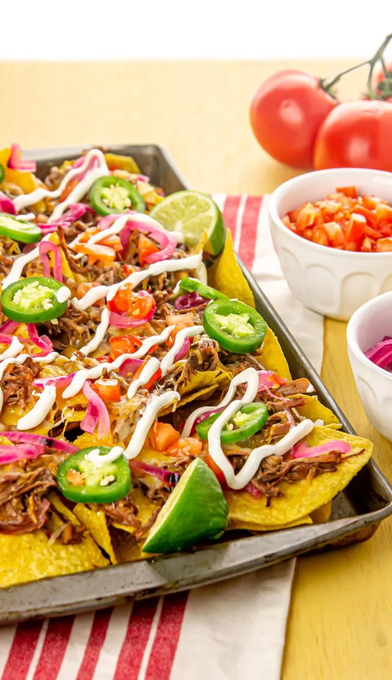 Tray of nachos topped with shredded beef, jalapeño slices, diced tomatoes, pickled onions, melted cheese, sour cream, and lime wedges.