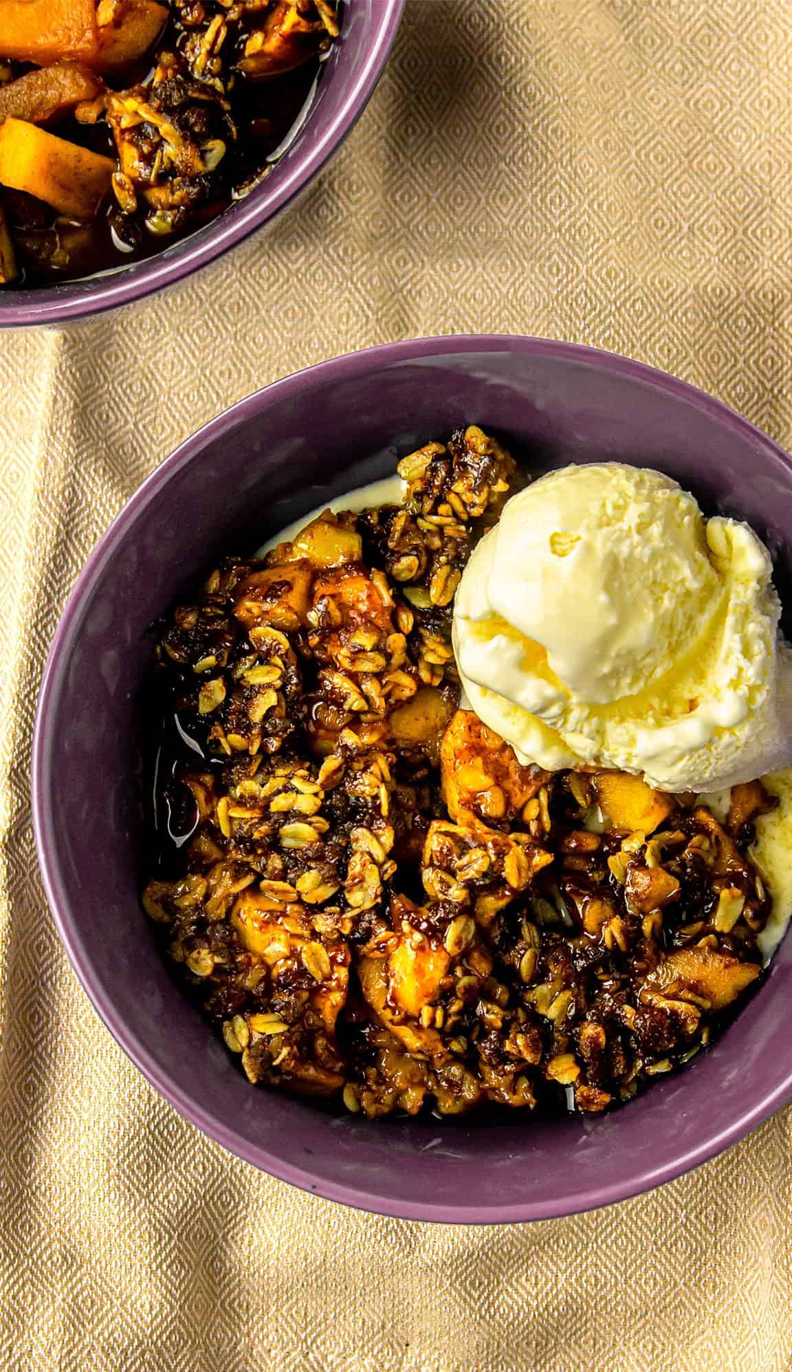Baked fruit crisp with oat topping served in purple bowl, topped with scoop of vanilla ice cream on beige patterned cloth.