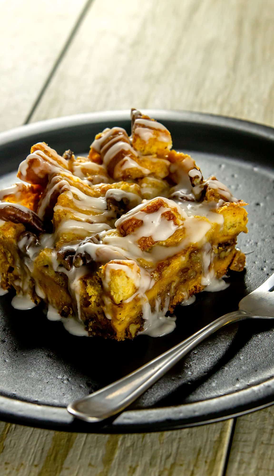 Cinnamon roll bread pudding square drizzled with white icing on a black plate with a fork.