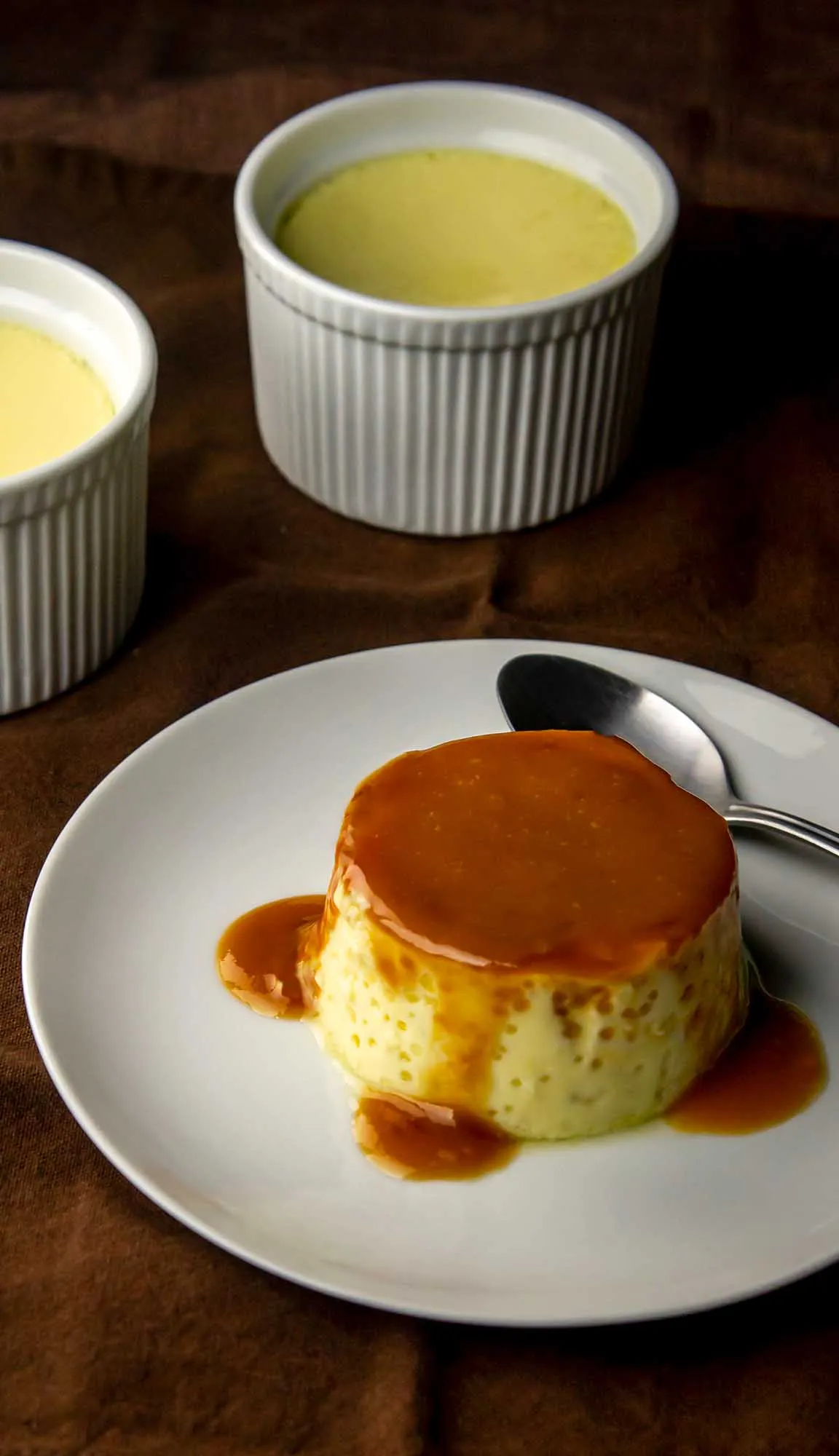 Caramel flan on a white plate with caramel sauce dripping down, spoon beside it, and two ramekins filled with custard in the background.