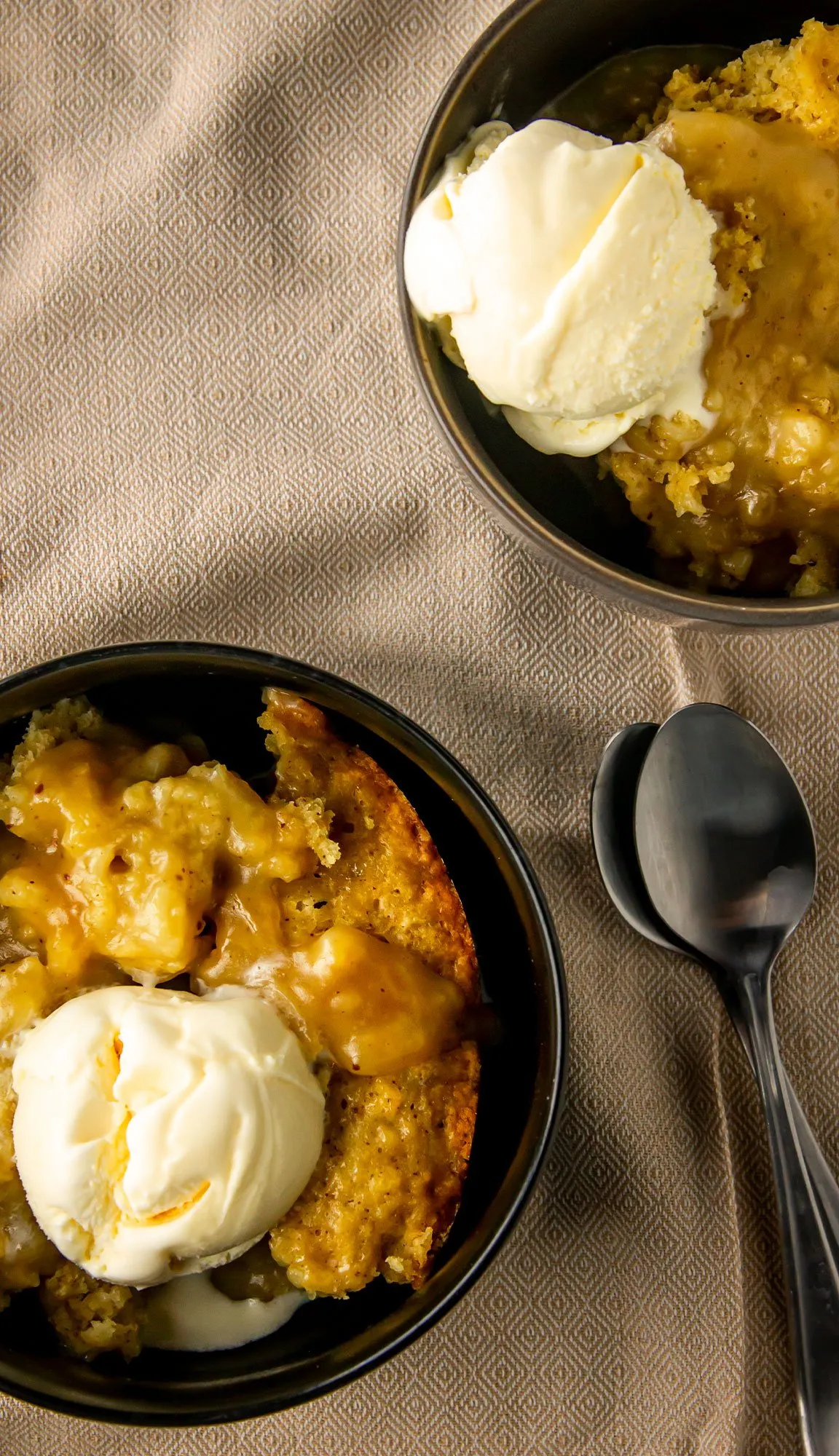Two black bowls with cobbler topped with vanilla ice cream on a textured beige cloth, next to two metal spoons.