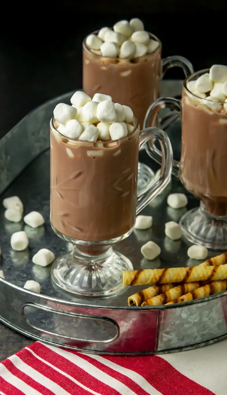 Three glass mugs of iced hot chocolate topped with marshmallows on a metal tray, with scattered marshmallows and wafer sticks nearby.