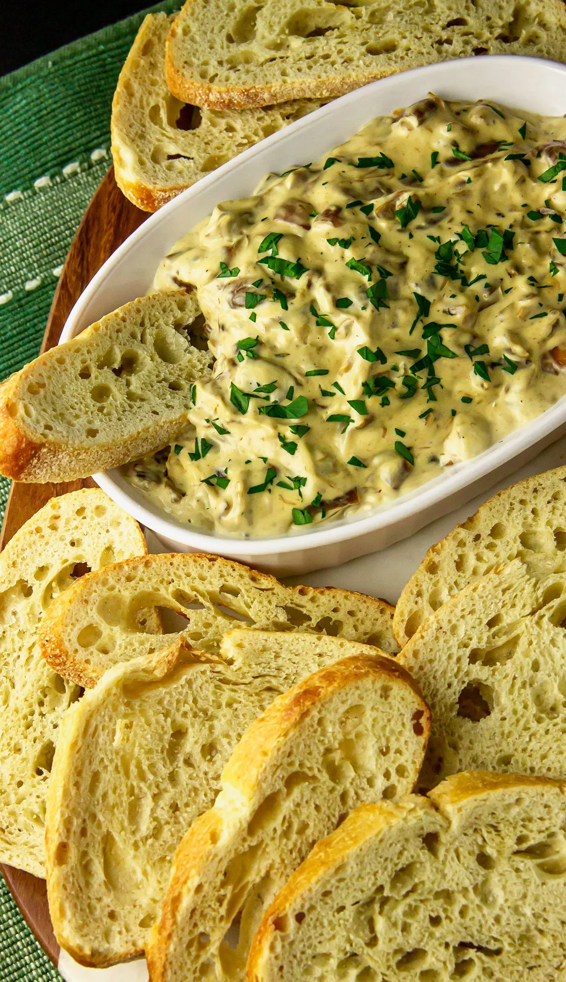 Creamy mushroom dip garnished with chopped herbs served with slices of crusty bread on a wooden board.