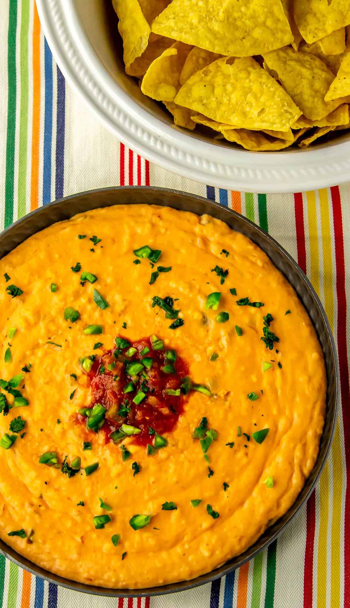 Cheese dip garnished with chopped jalapeños and parsley, served with a bowl of tortilla chips on a striped cloth background.