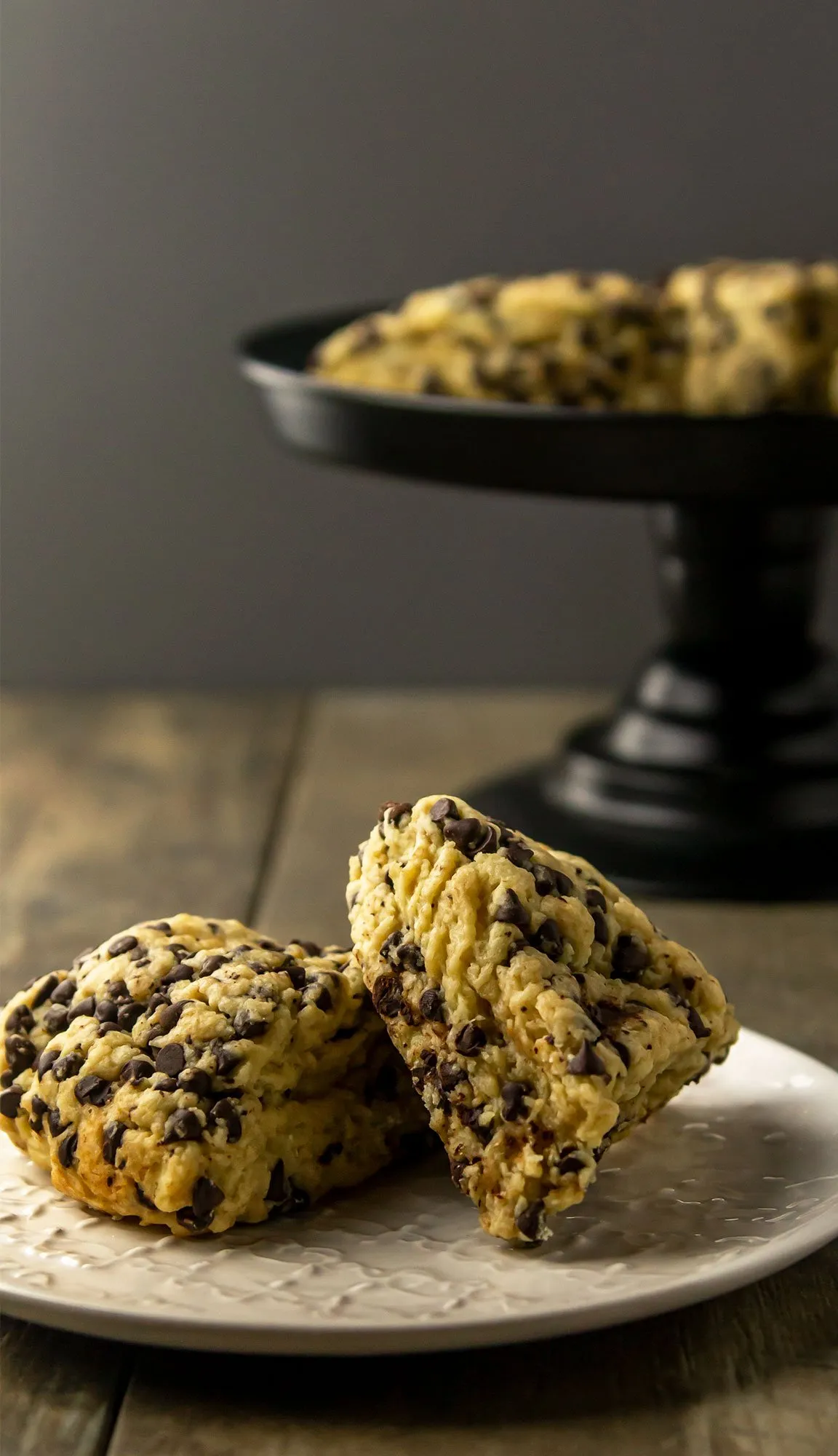 Two chocolate chip scones on a white plate with more scones on a black cake stand in the background.