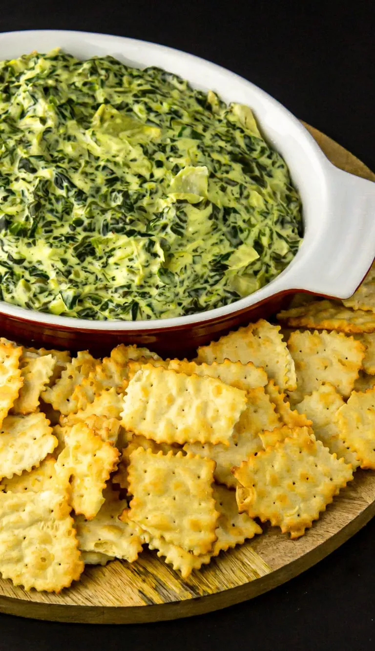 Creamy spinach artichoke dip in a white bowl served with golden square crackers on a wooden board.