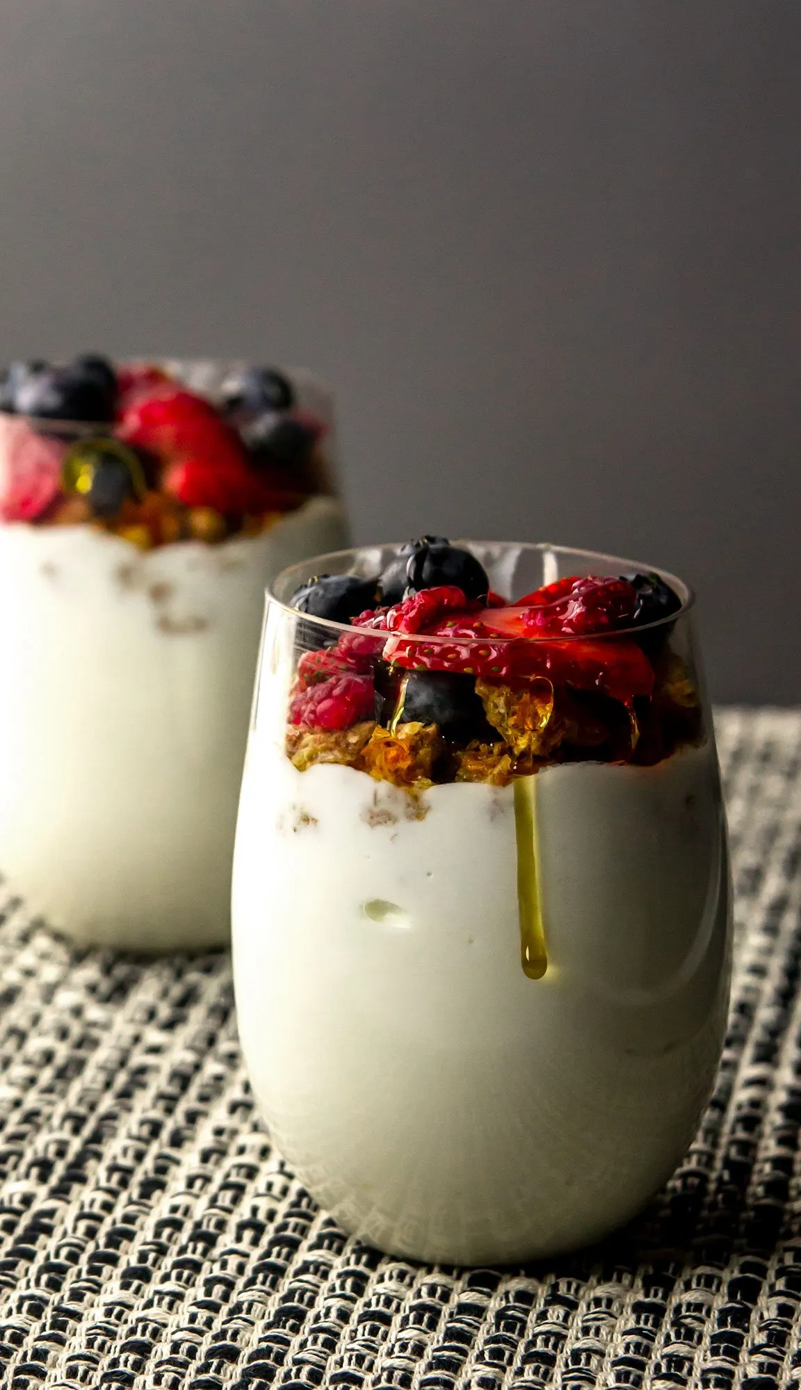 Two glasses of yogurt parfait topped with granola, blueberries, raspberries, and honey on a textured mat with a gray background.