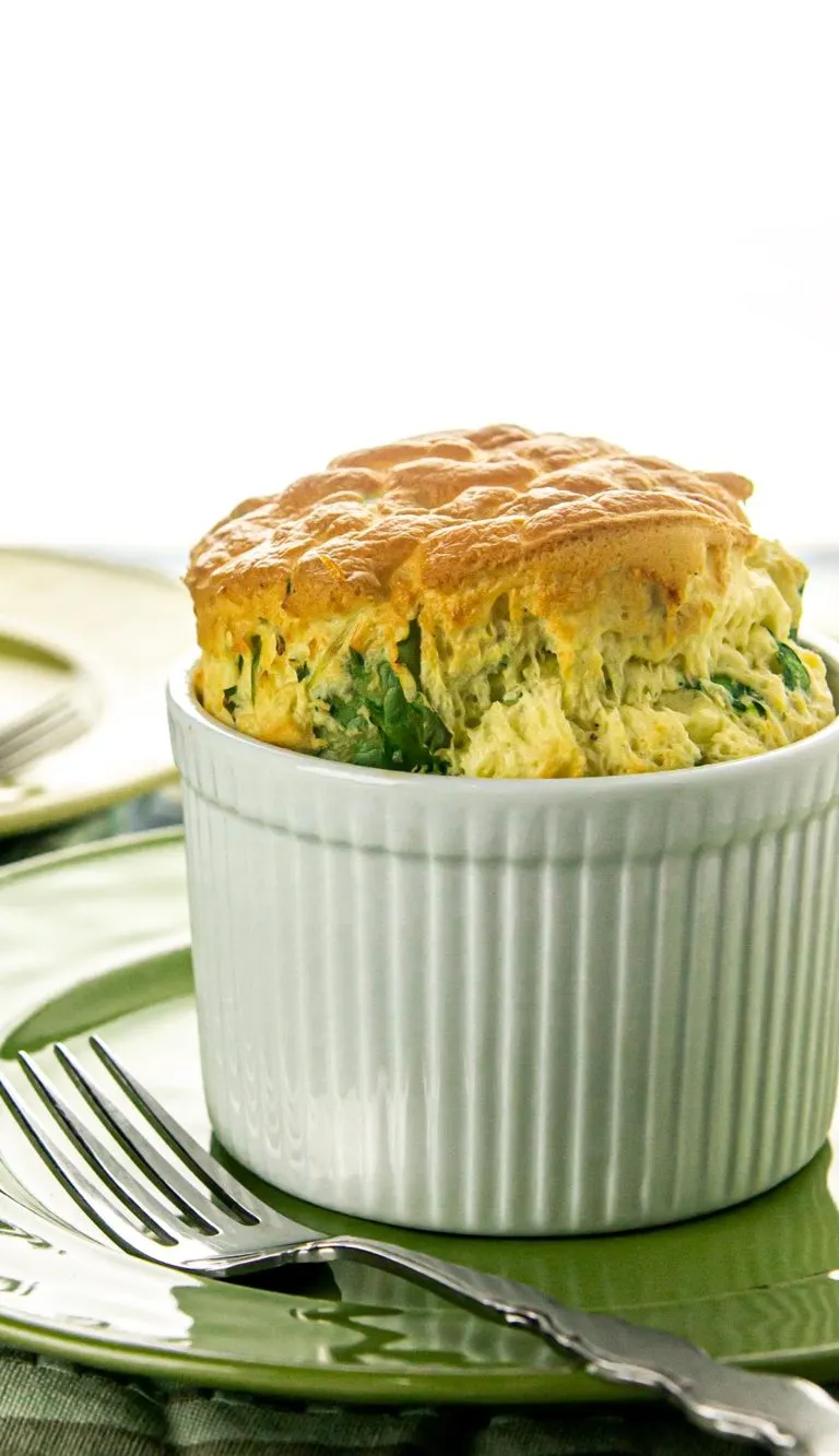 Fluffy spinach and cheese soufflé rising in a white ceramic ramekin on a green plate with a fork.