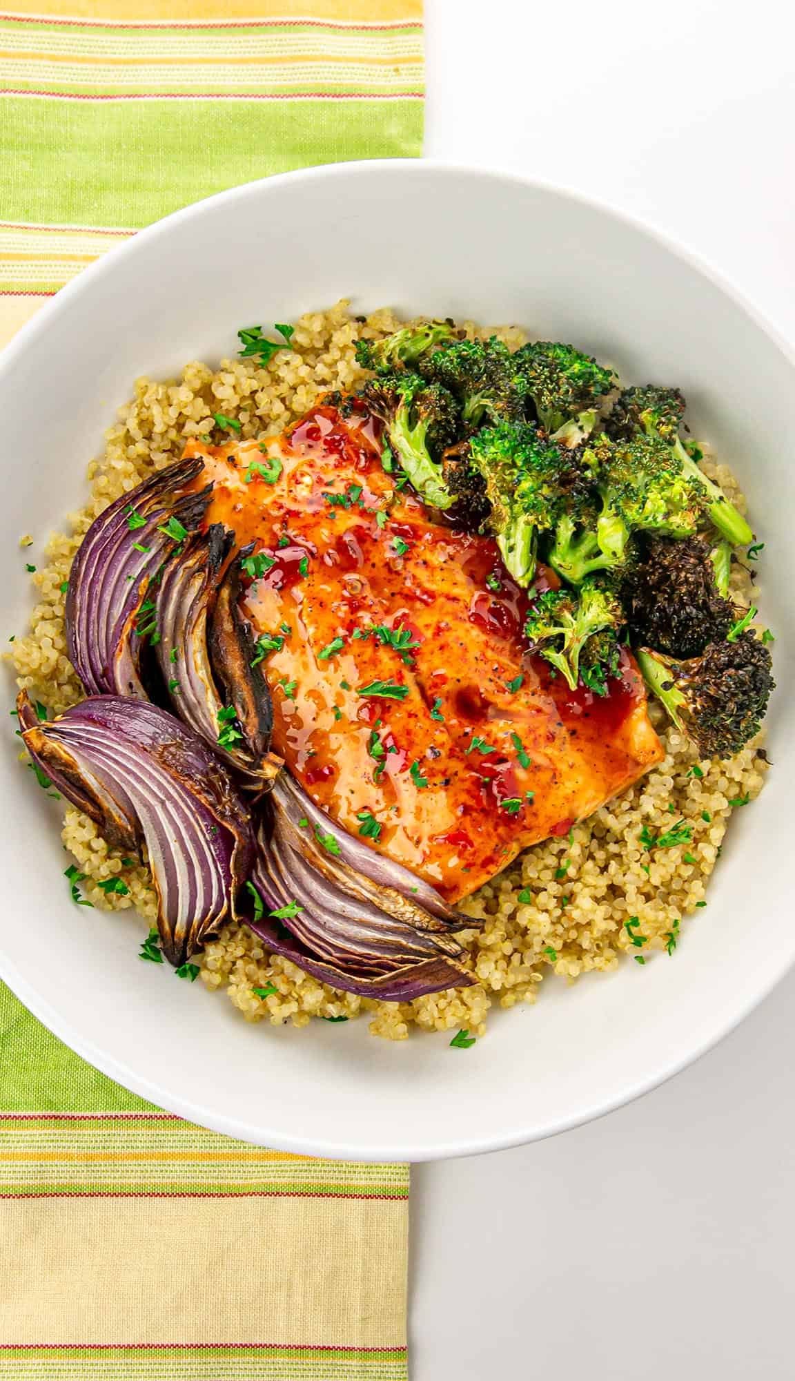 Grilled salmon with roasted red onions, broccoli, and quinoa in a white bowl on a striped green and yellow napkin.
