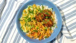 White rice bowl with three meatballs, sliced cucumber, shredded carrots, drizzled with orange sauce, and garnished with chopped herbs on a blue-striped cloth.