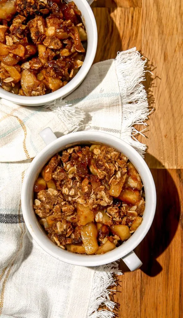 Two white bowls filled with apple crumble on a fringed cloth over a wooden surface.